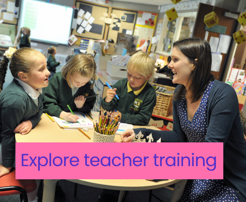A teacher at a round table with 3 primary school children in green jumpers. One is talking to the teacher, the other two are working on paper worksheets with a green pencil and blue pencil. In the background is a classroom with more children visible.